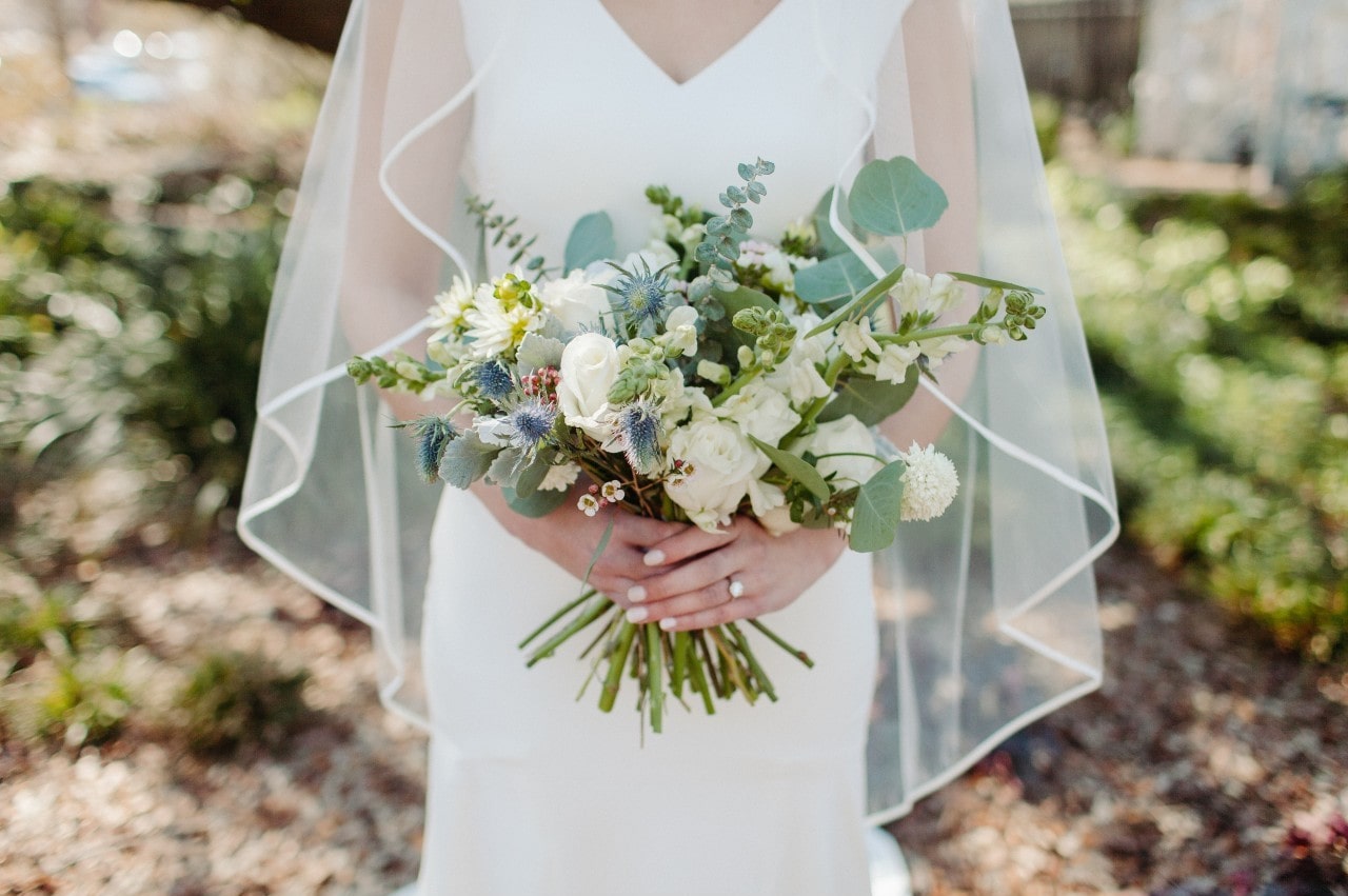 A bride with a veil holds her bouquet while standing outside.