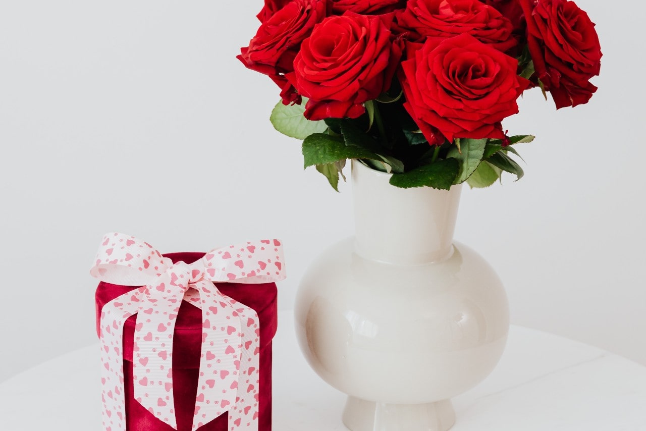 A pink round gift box with white and pink ribbon stands near the white vase with several red roses against a white background.