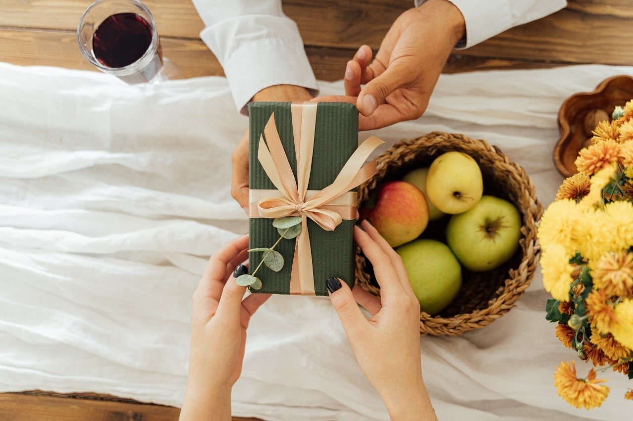 A close-up view of two people exchanging a green wrapped gift over a table with apples, flowers, and a white tablecloth.