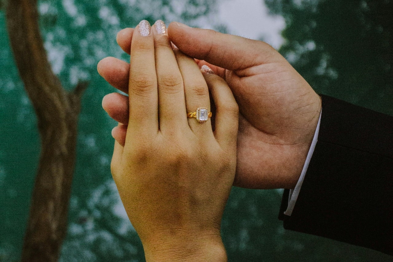 A close up of a man’s hand holding a woman’s hand, showcasing yellow gold halo engagement ring with emerald cut center diamond.