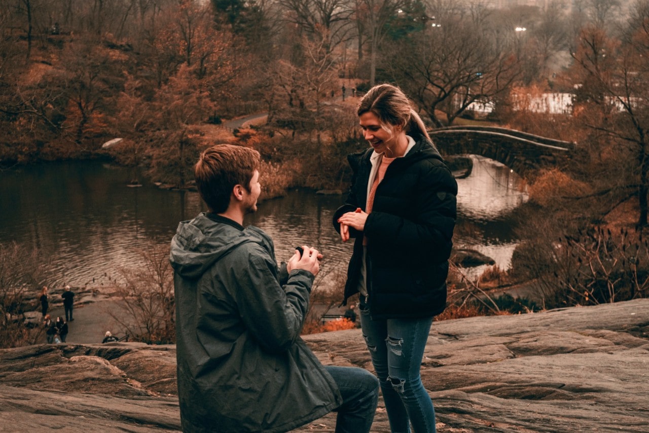 A man proposes to his girlfriend on a hill overlooking a fall lake.