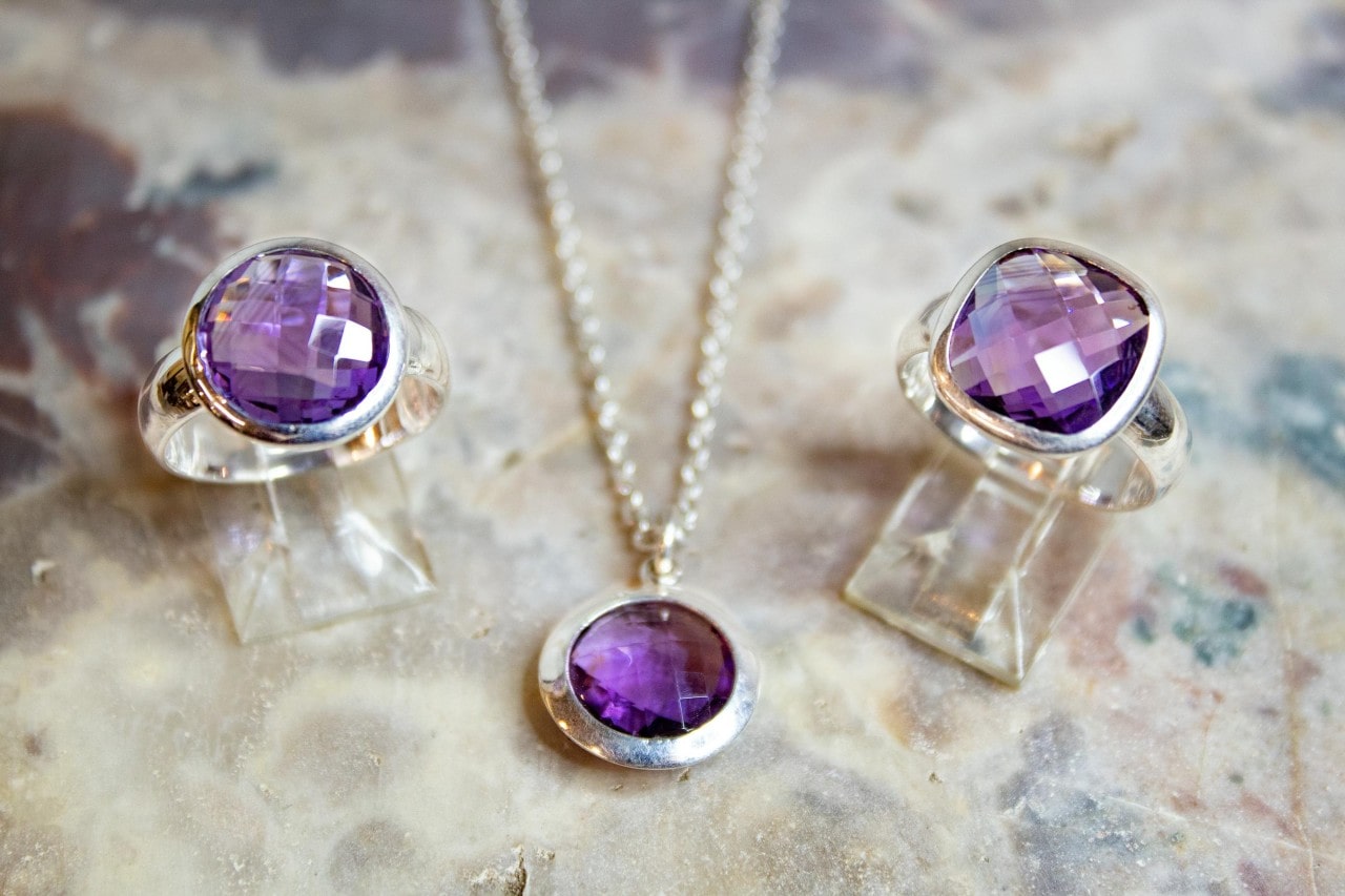 A faceted amethyst jewelry set on a marble surface, featuring a bezel set solitaire necklace and two fashion rings.