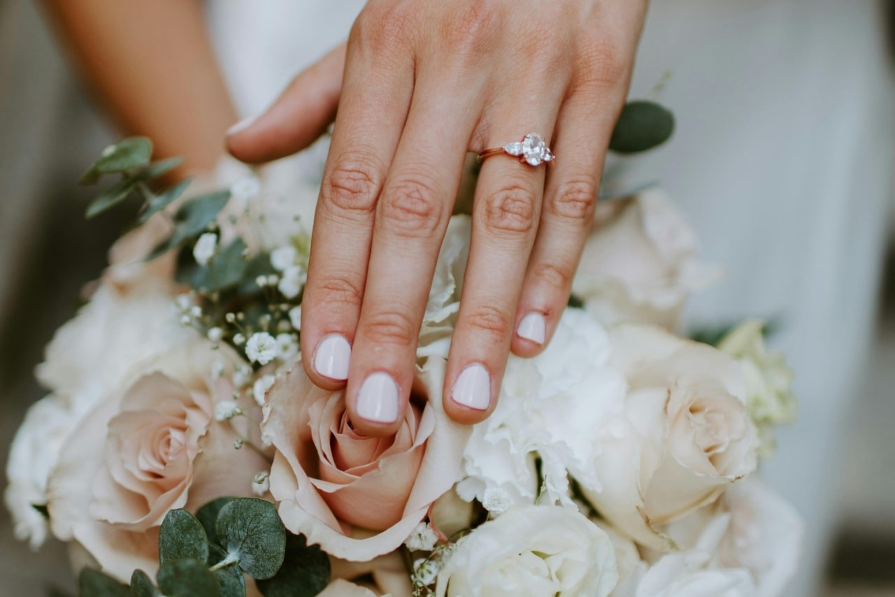 A close-up of a woman&rsquo;s hand showing an engagement ring while resting on a bouquet of roses and greenery.