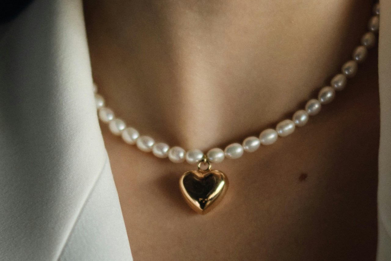 A close up image of a woman’s neckline adorned with a pearl necklace featuring a heart pendant