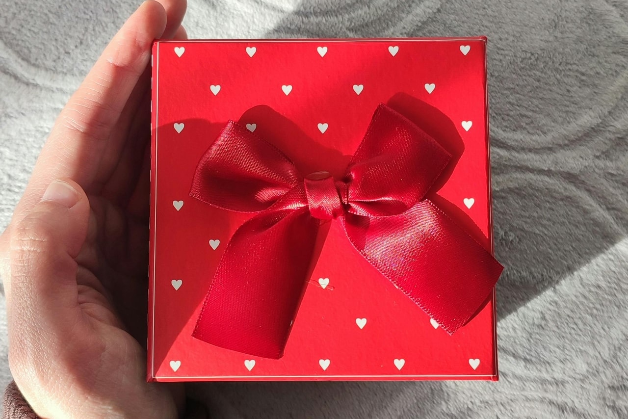A close-up of a cute red gift box with white flowers resting atop a soft blanket next to a man’s open hand.