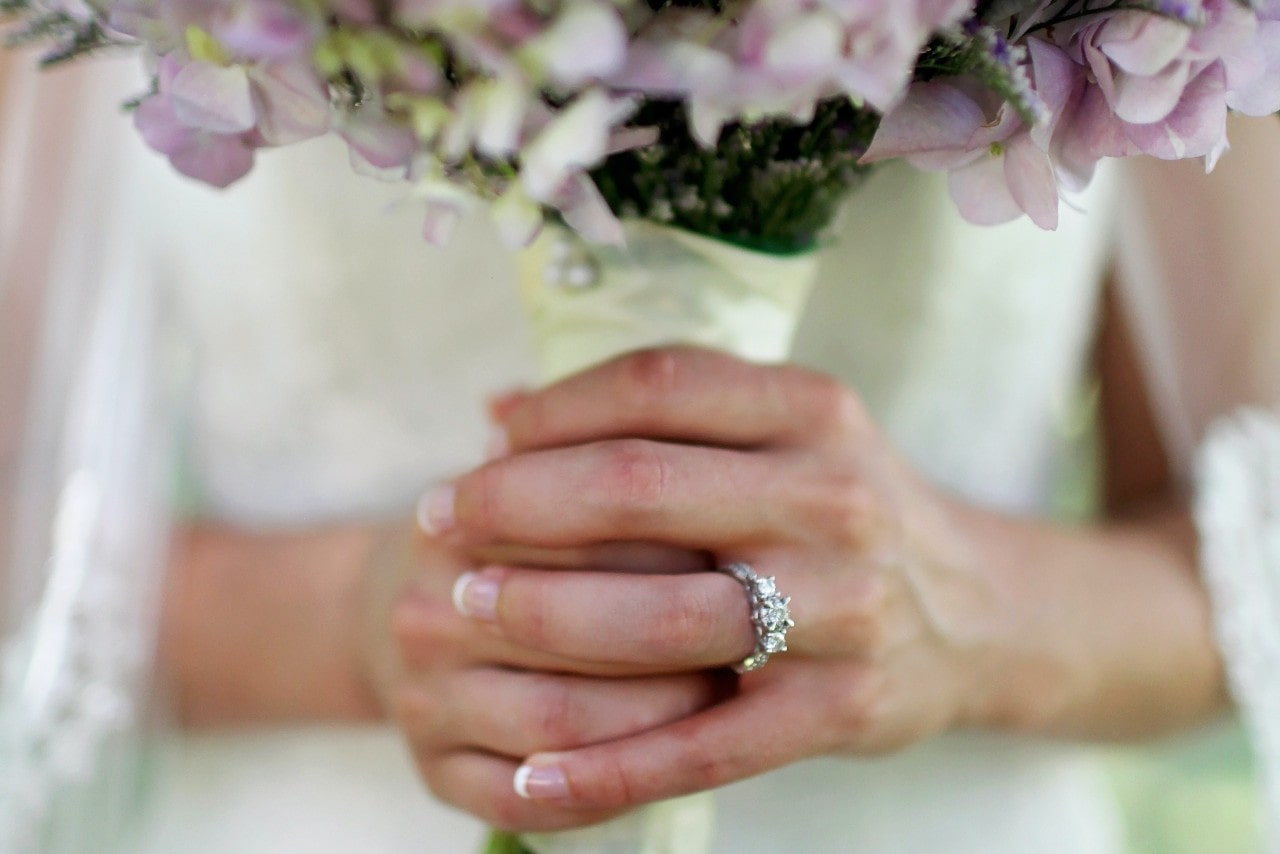 Close up of bride’s hand holding a bouquet and wearing a three stone, round cut engagement ring