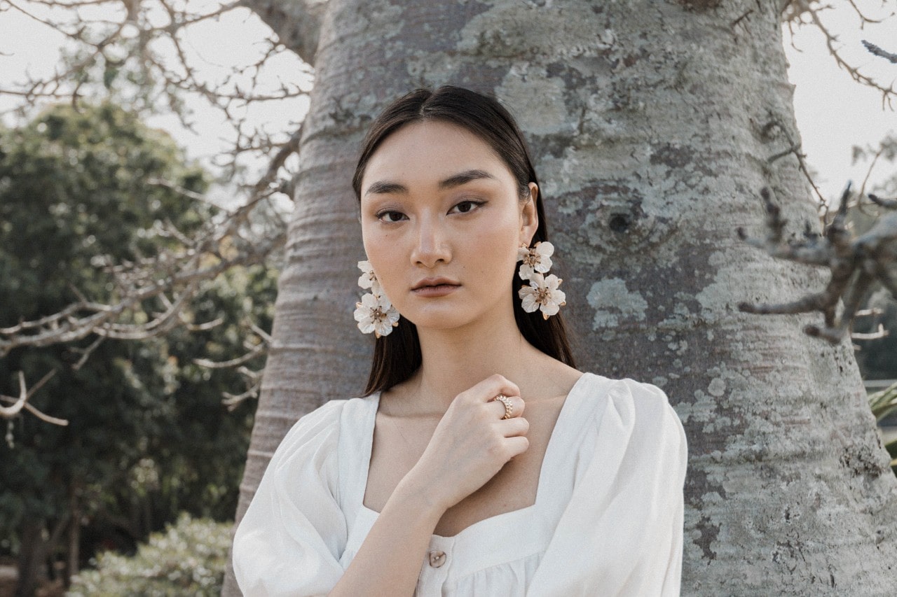 a woman standing in front of a tree wearing white and pair of floral earrings