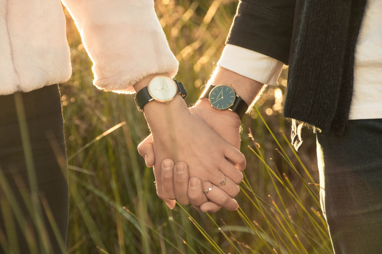 A couple holding hands walks through a field while wearing watches