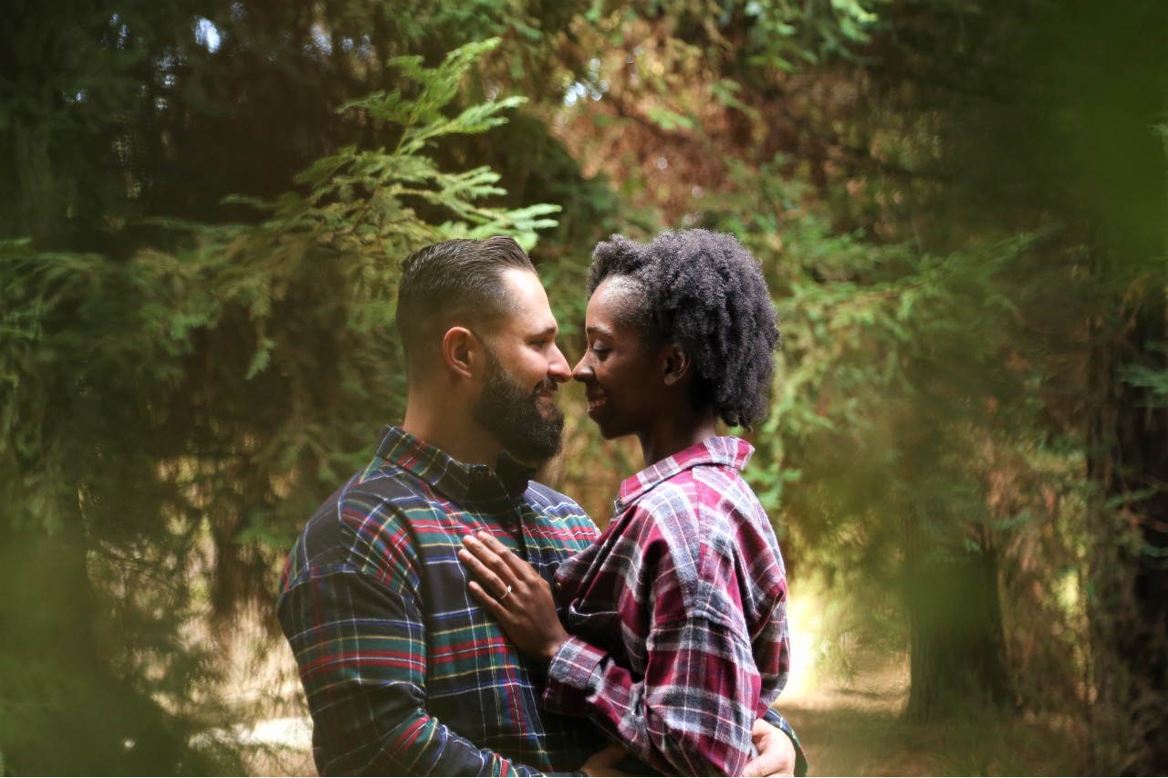 A couple cuddles wearing flannel shirts embracing each other while being surrounded by trees in the autumn setting.