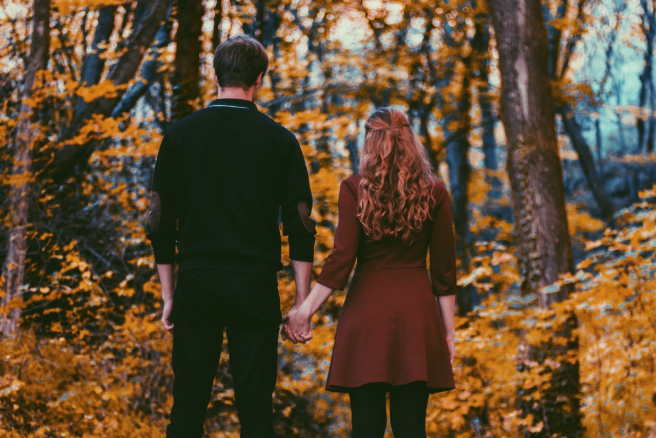 A couple hold hands while standing in front of autumn forest scenery.