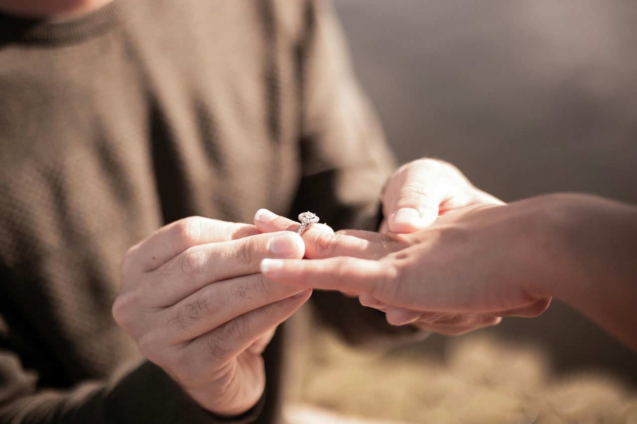 A person putting an engagement ring on another’s finger during a proposal, with natural lighting and a blurred background.