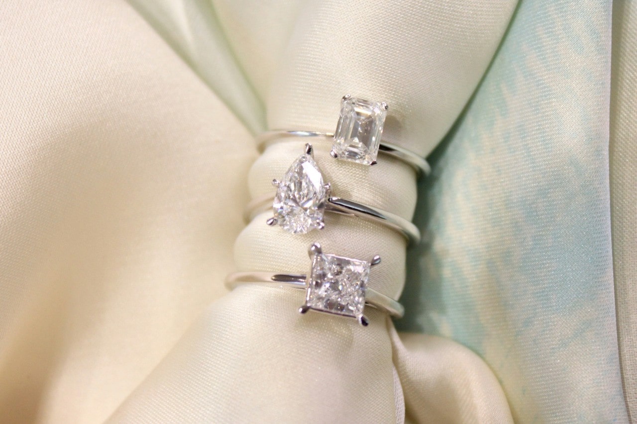 Three solitaire diamond engagement rings with emerald, pear, and princess cuts displayed elegantly on satin fabric