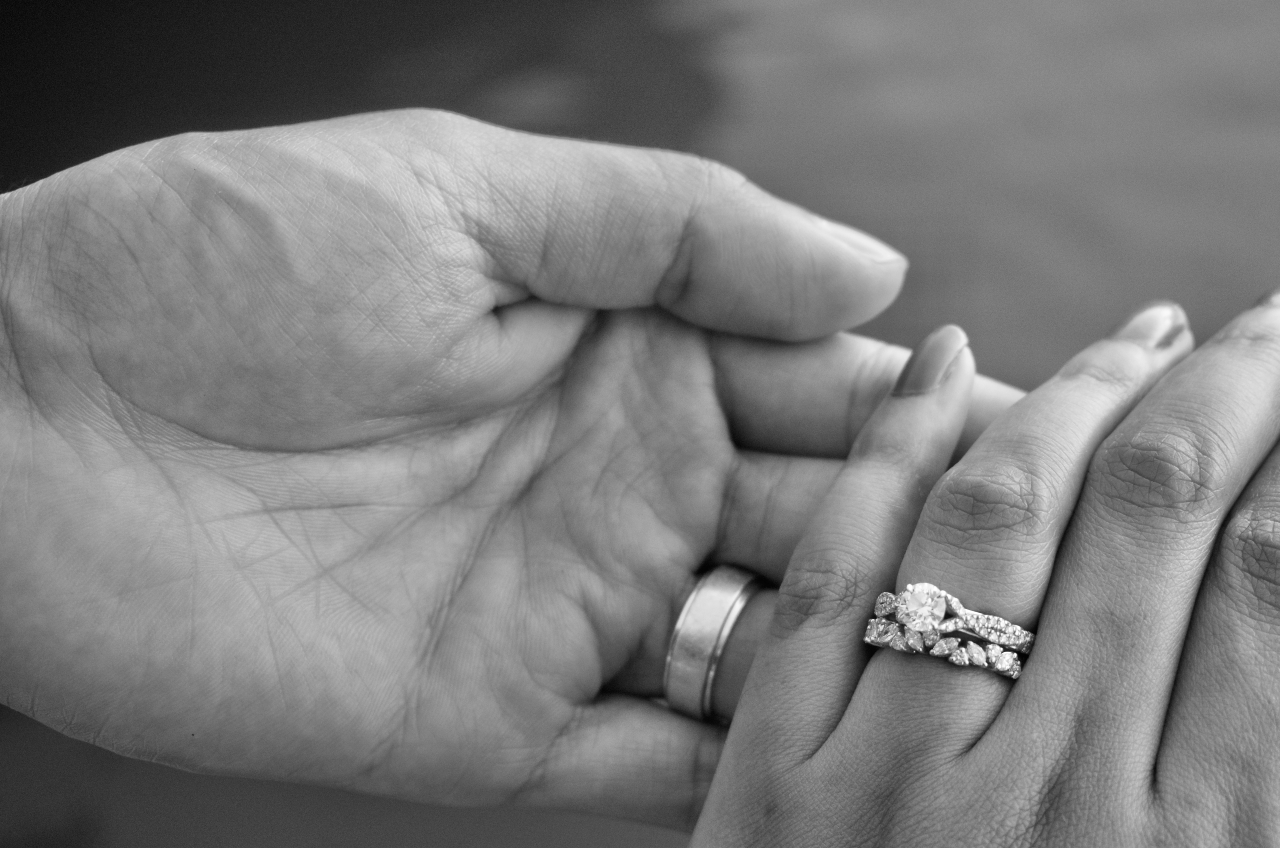 A black and white close-up of two hands holding each other, showcasing a wedding and engagement ring set.