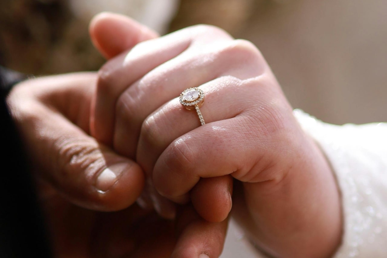 A close-up of two hands together, highlighting a halo diamond engagement ring with a pavé band on a lady's finger.