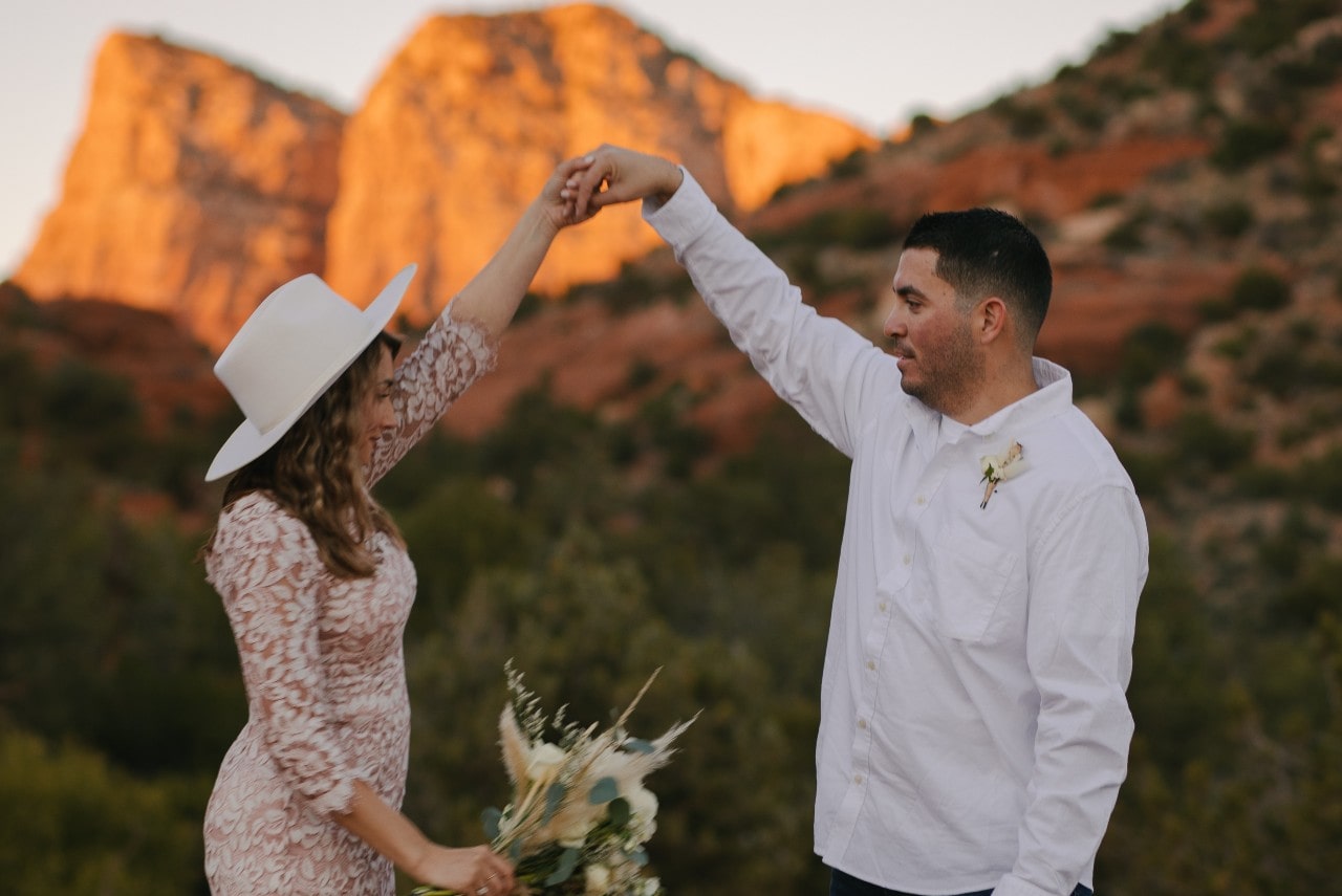 Husband and wife holding hands with rock formations and mountains in the background. The bride is holding a bouquet and wearing a white felt hat.
