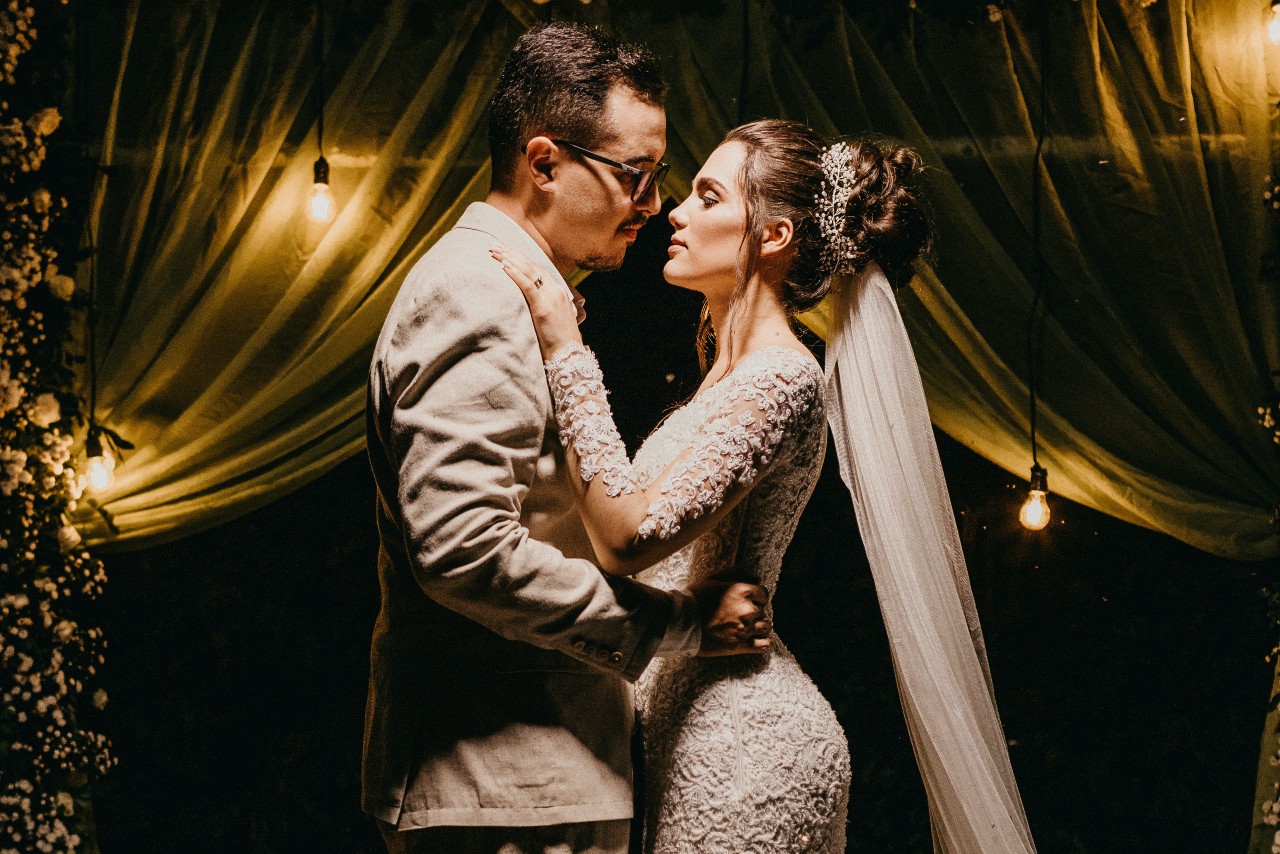 Groom dressed in a white tux dances with the bride in a lacy, long sleeve dress under lights and flowers.