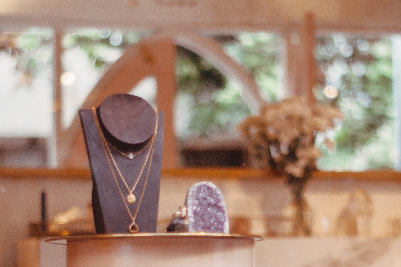 Elegant jewelry display with layered gold necklaces on a stand, set in a softly lit boutique, with blurred floral accents in the background.