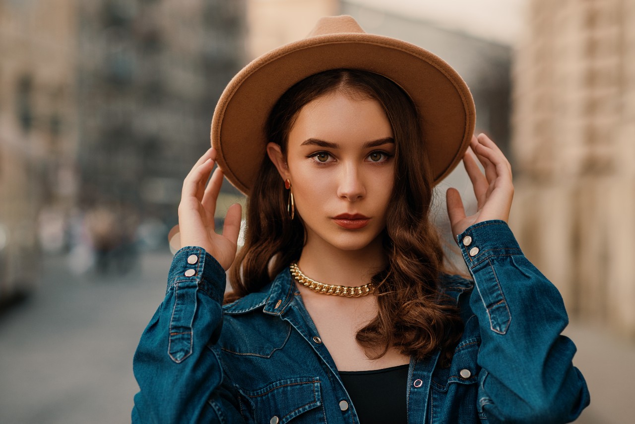 A woman in a denim jacket and tan hat stands on a city street, touching her hat, with gold hoop earrings catching the light.