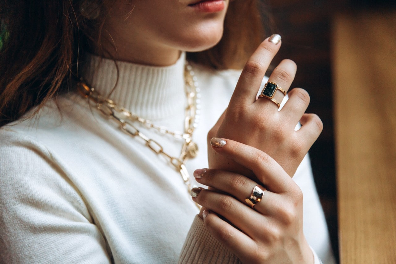 A woman with a white sweater, layered gold chain necklaces, and two contemporary style rings.