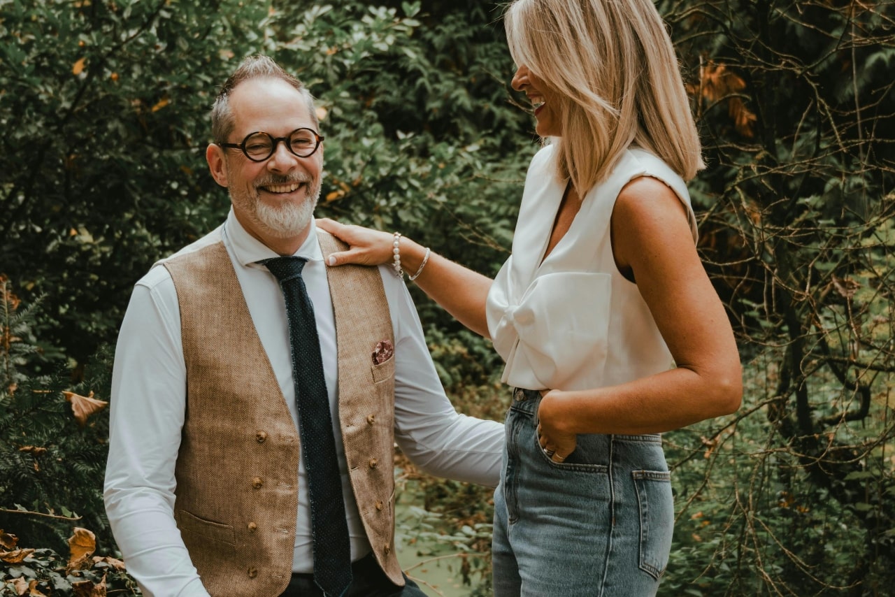 A couple laugh together as they pose in the woods for a picture during a family event