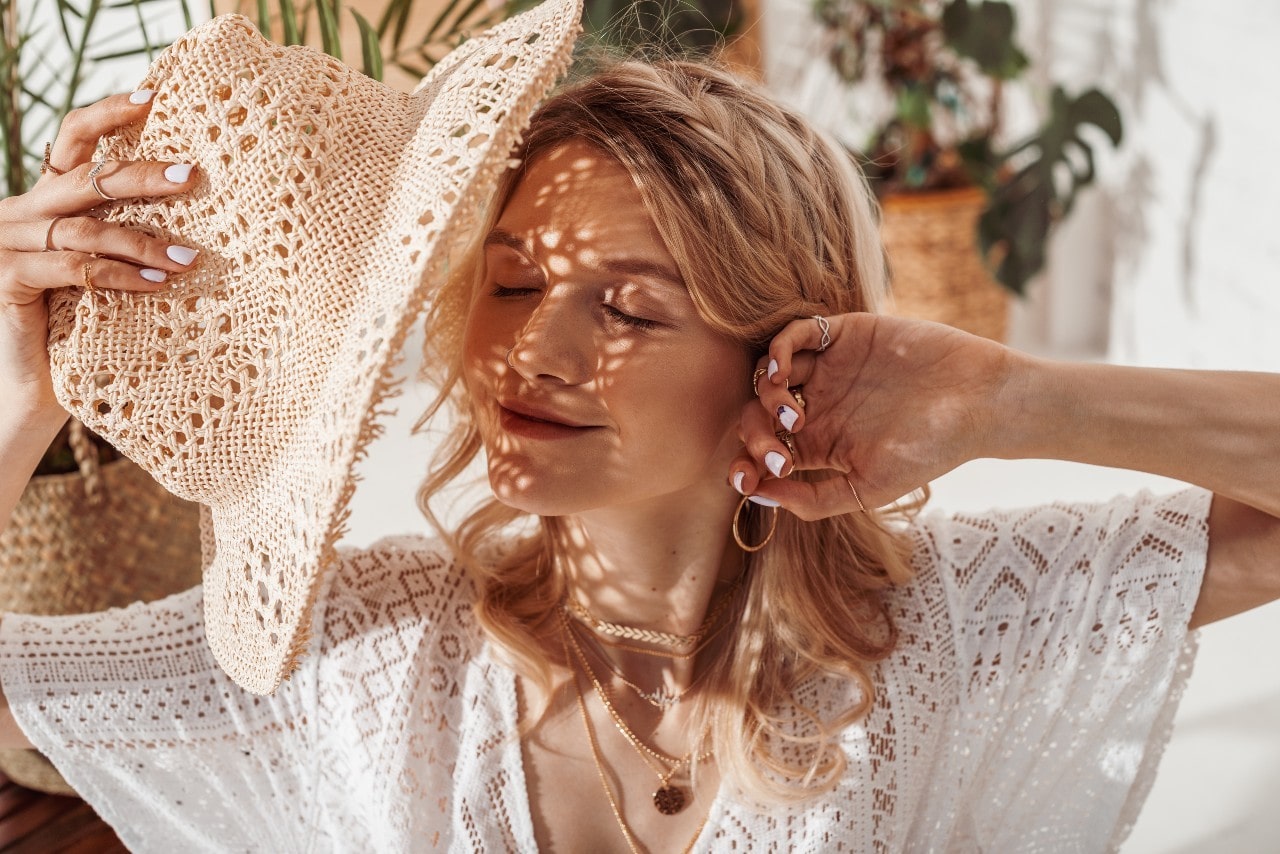 A woman hides her face with her straw hat while wearing a lacy top with jewelry.