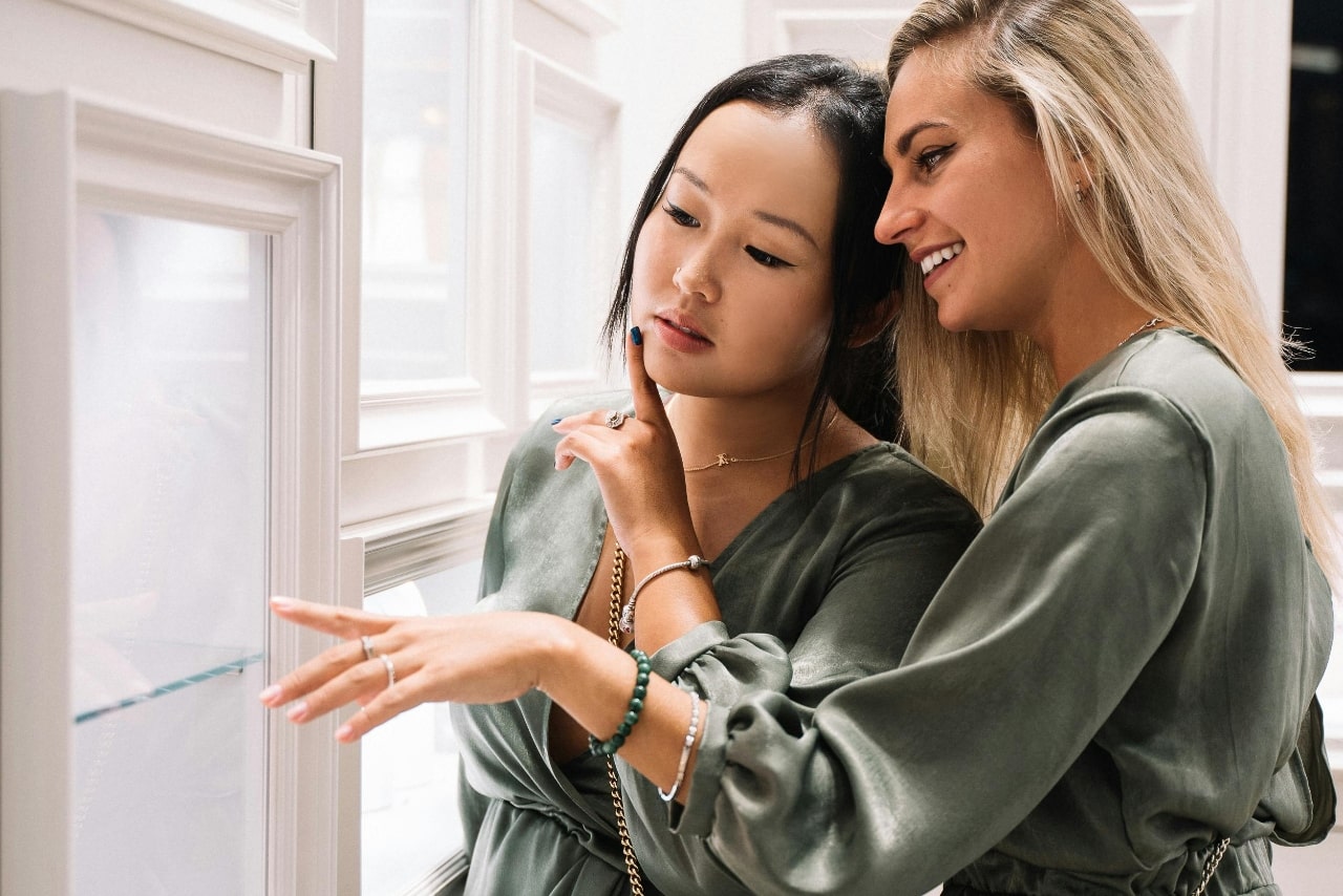 Two women in green satin blouses admire jewelry in a display case.
