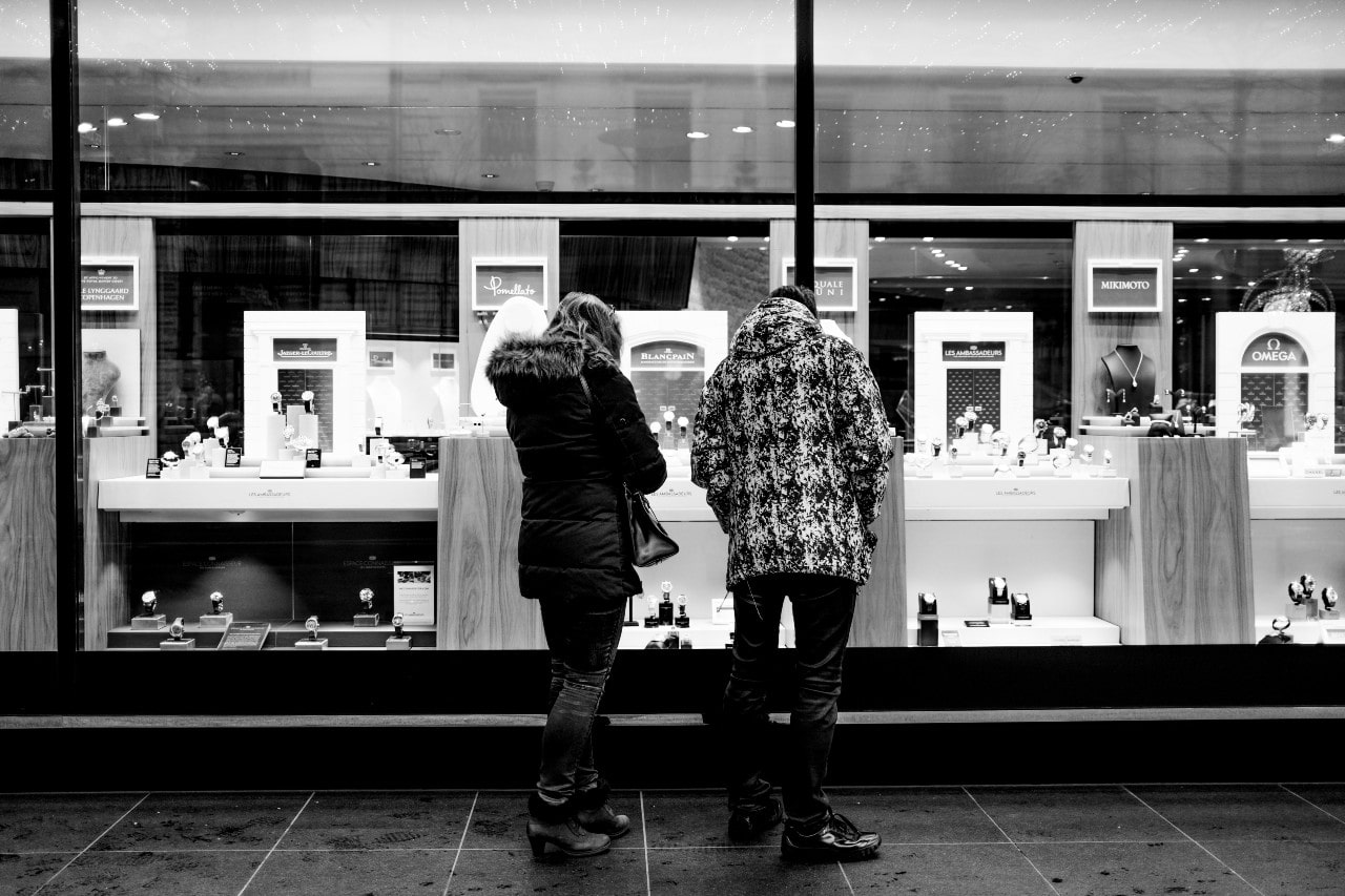 A black-and-white photo shows two people in winter jackets gazing into a well-lit jewelry store window.