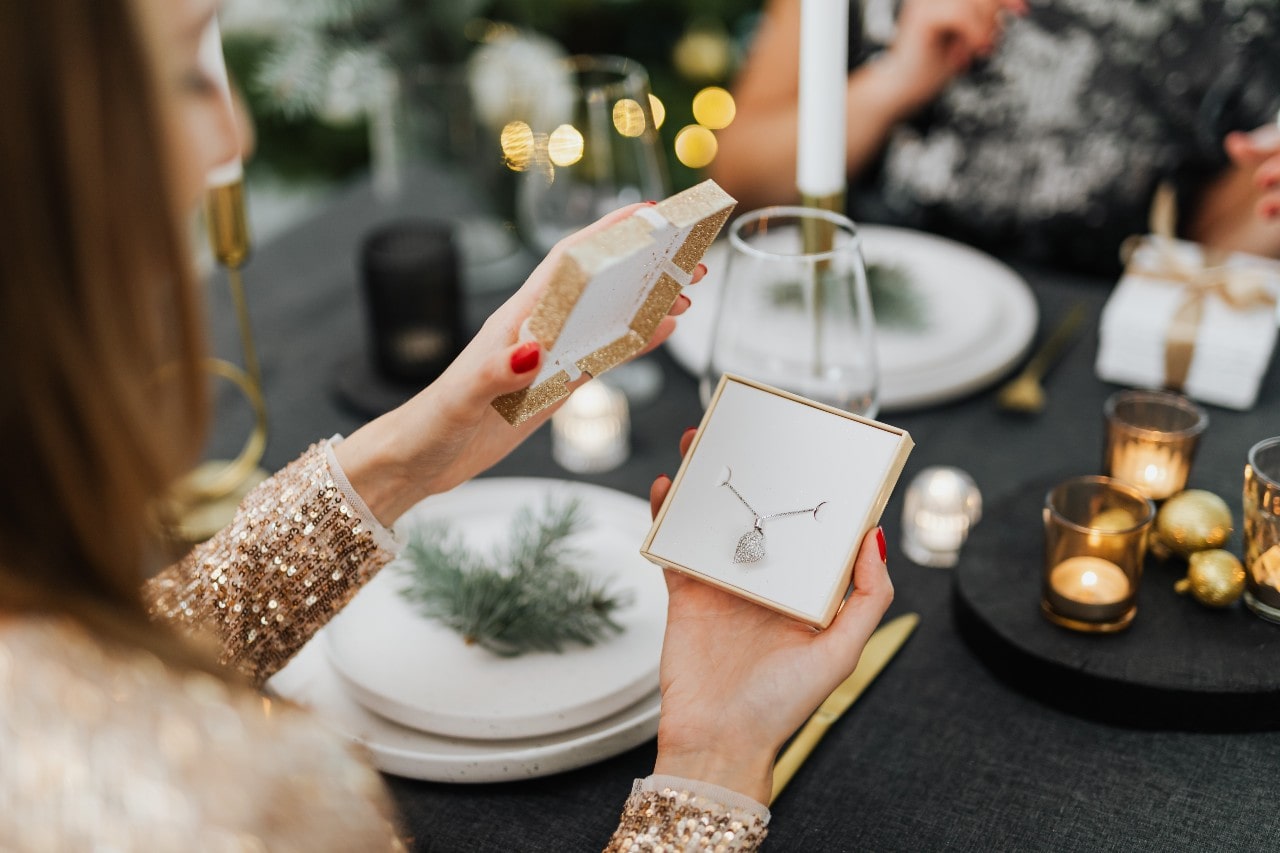 A woman in a sequined dress opens a glittery gift box with a silver heart-shaped pendant necklace, while sitting at a candlelit dinner table.