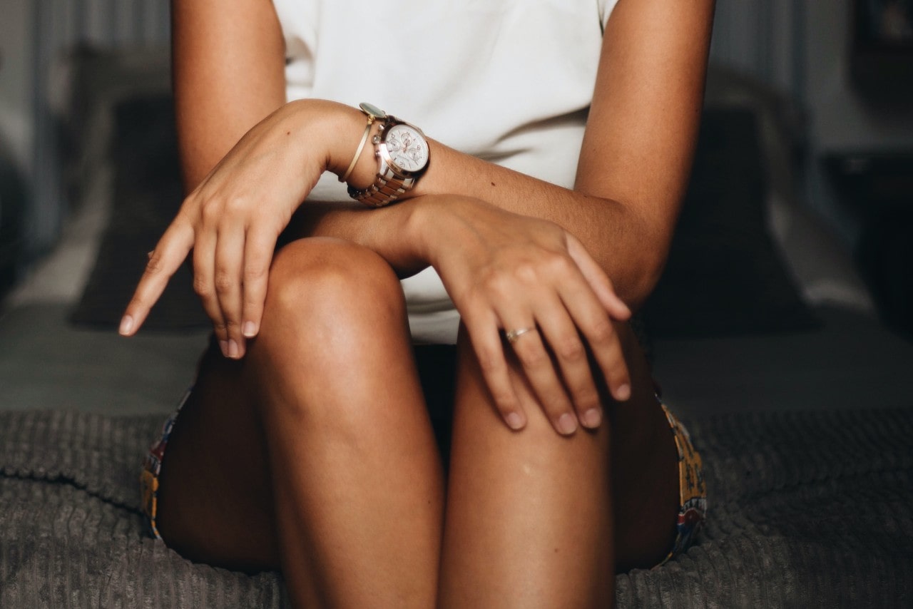 A woman sitting on the end of a bed with wrists crossed, wearing a white and gold watch