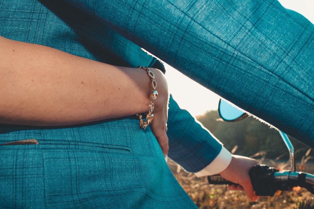 A man in a suit driving a motorcycle while a woman wearing a Fana bracelet wraps her arms around his waist.