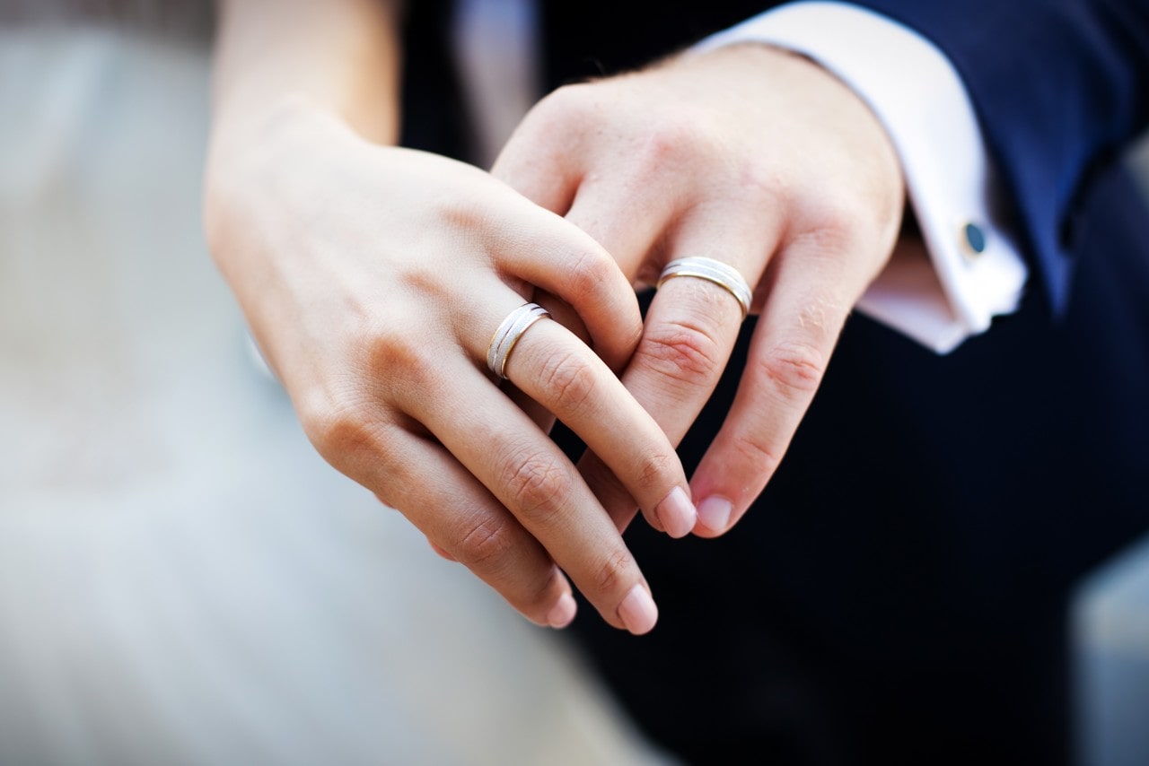 A pair of intertwined hands wearing matching white gold wedding bands.