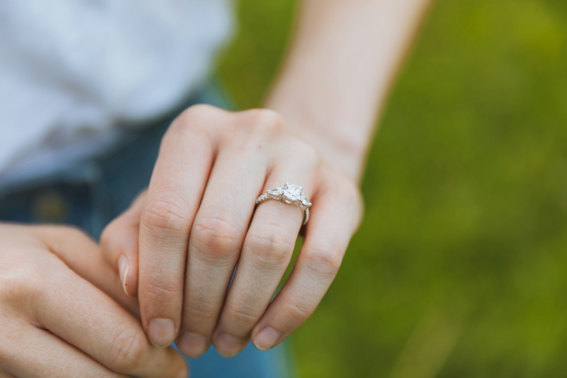 A close up of a three stone engagement ring on a woman’s hand against a blurry grass