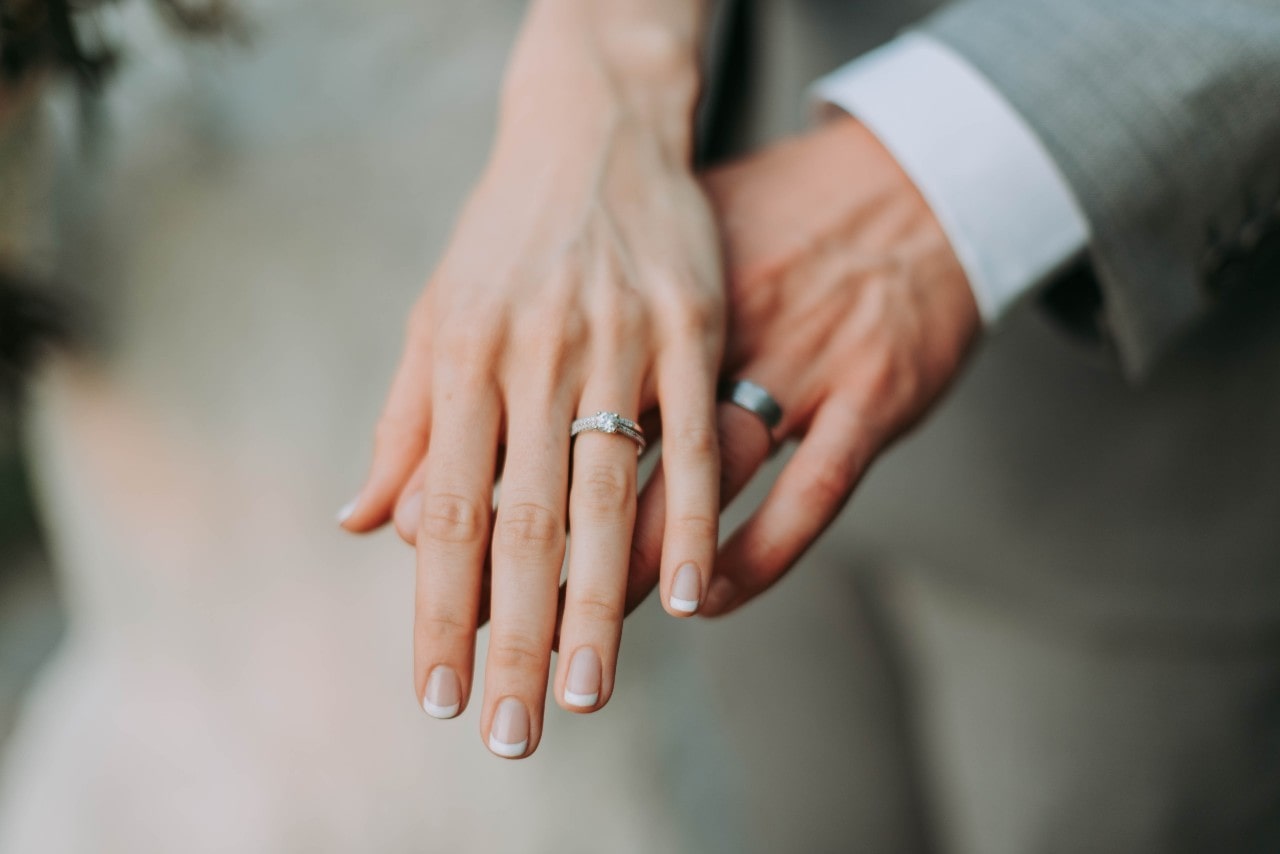 A close-up of a bride and groom's hands showing their wedding bands, with the bride wearing a French manicure.