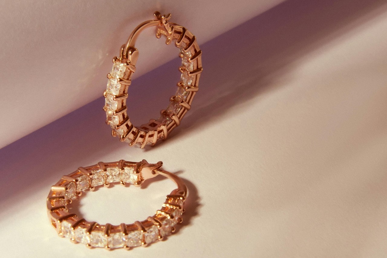 A close-up of an elegant pair of rose gold diamond hoop earrings on a beige backdrop.