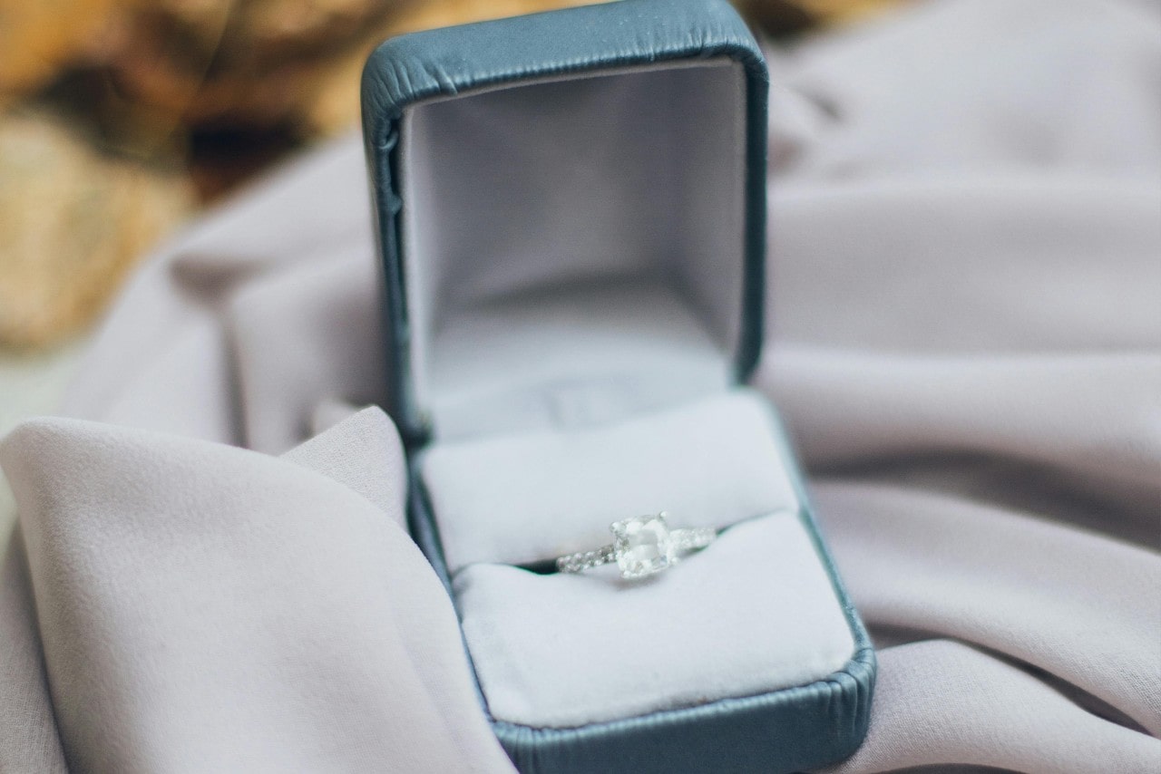 A white gold side stone engagement ring displayed inside its box, on top of white fabric. A white gold side stone engagement ring displayed inside its box, on top of white fabric.