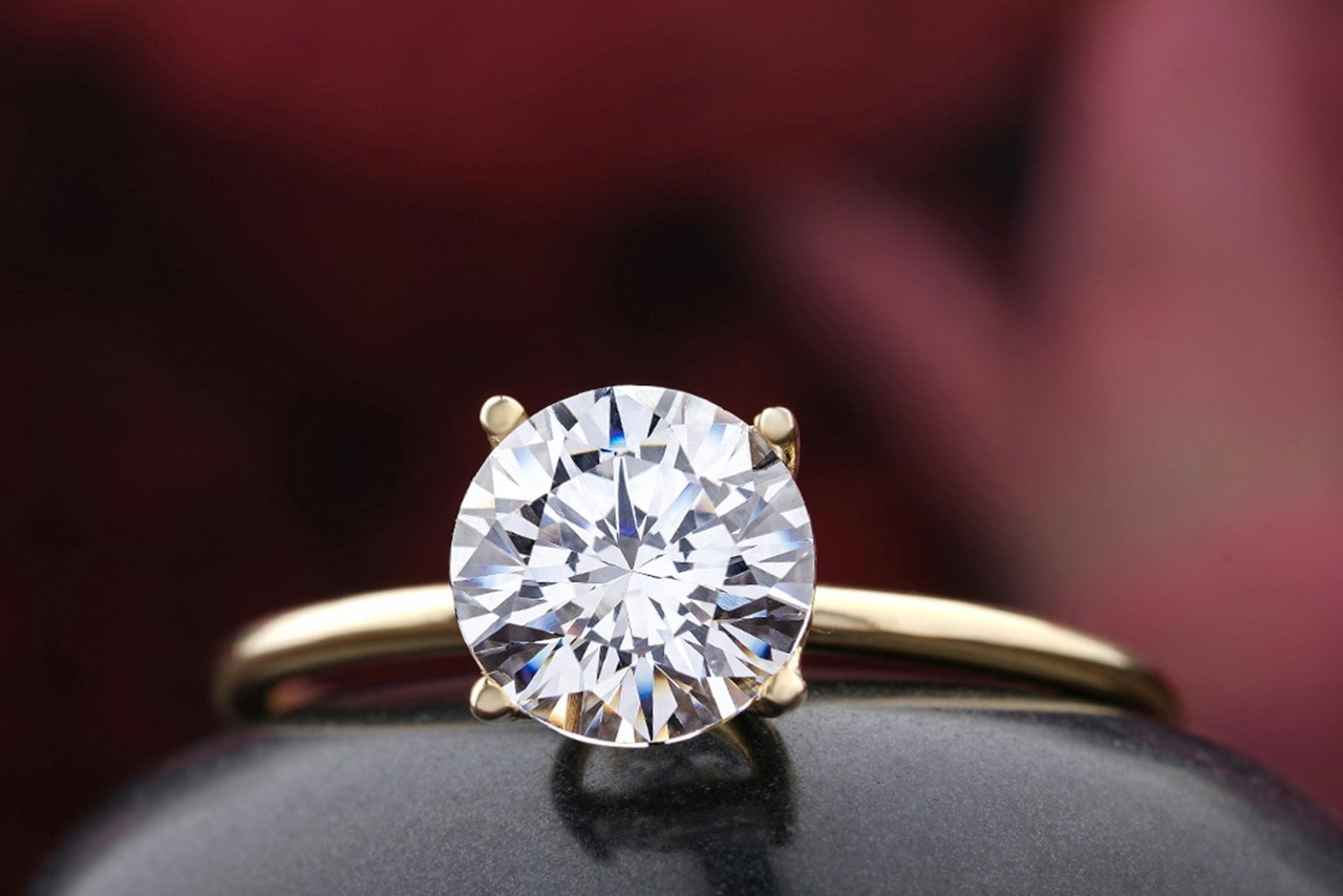 A close up of a round cut diamond solitaire with yellow gold polished band against a blurry dark red background A close up of a round cut diamond solitaire with yellow gold polished band against a blurry dark red background