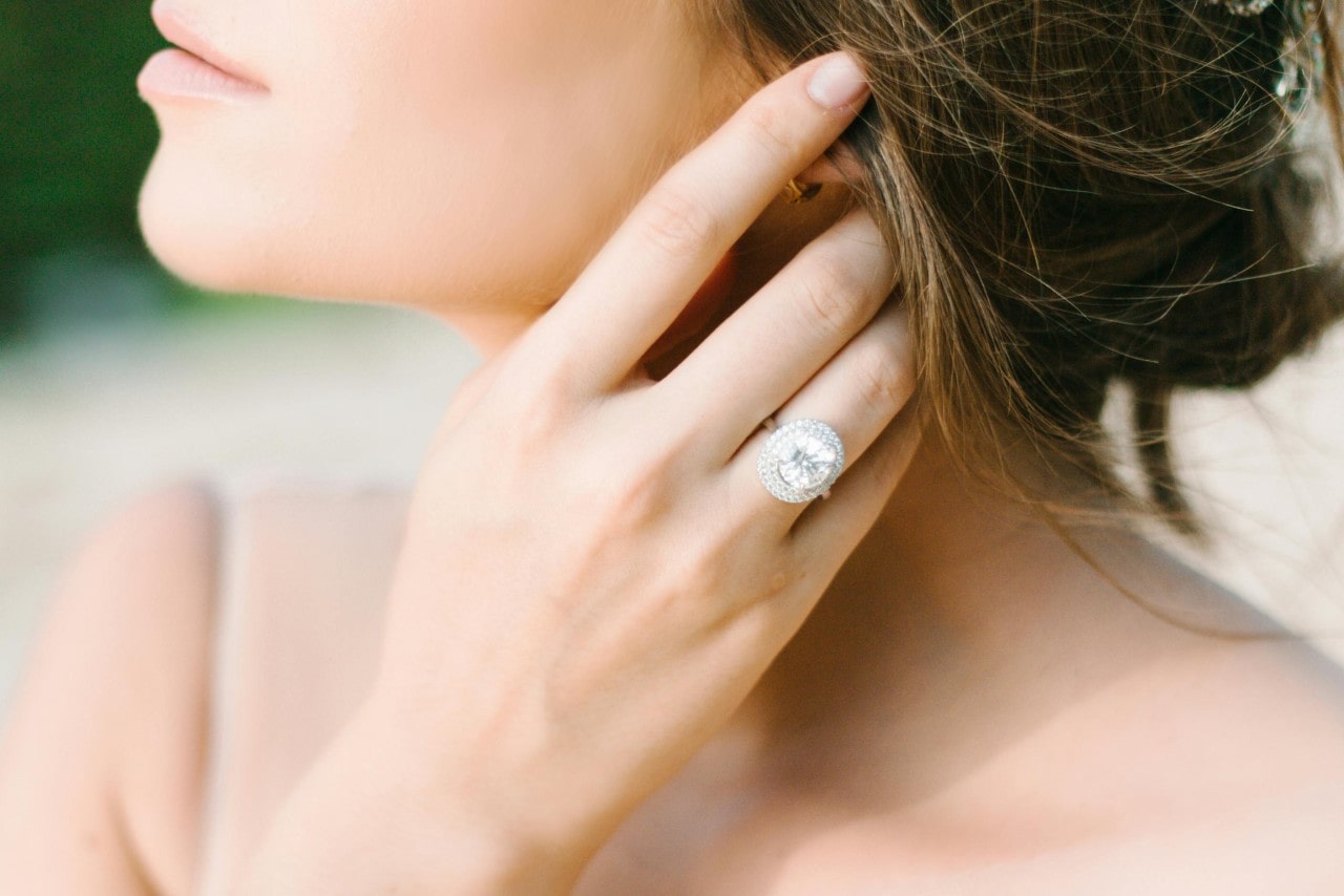 A close-up of a bride tucking a lock of hair behind her ear, a dramatic white gold halo diamond engagement ring on her finger. A close-up of a bride tucking a lock of hair behind her ear, a dramatic white gold halo diamond engagement ring on her finger.