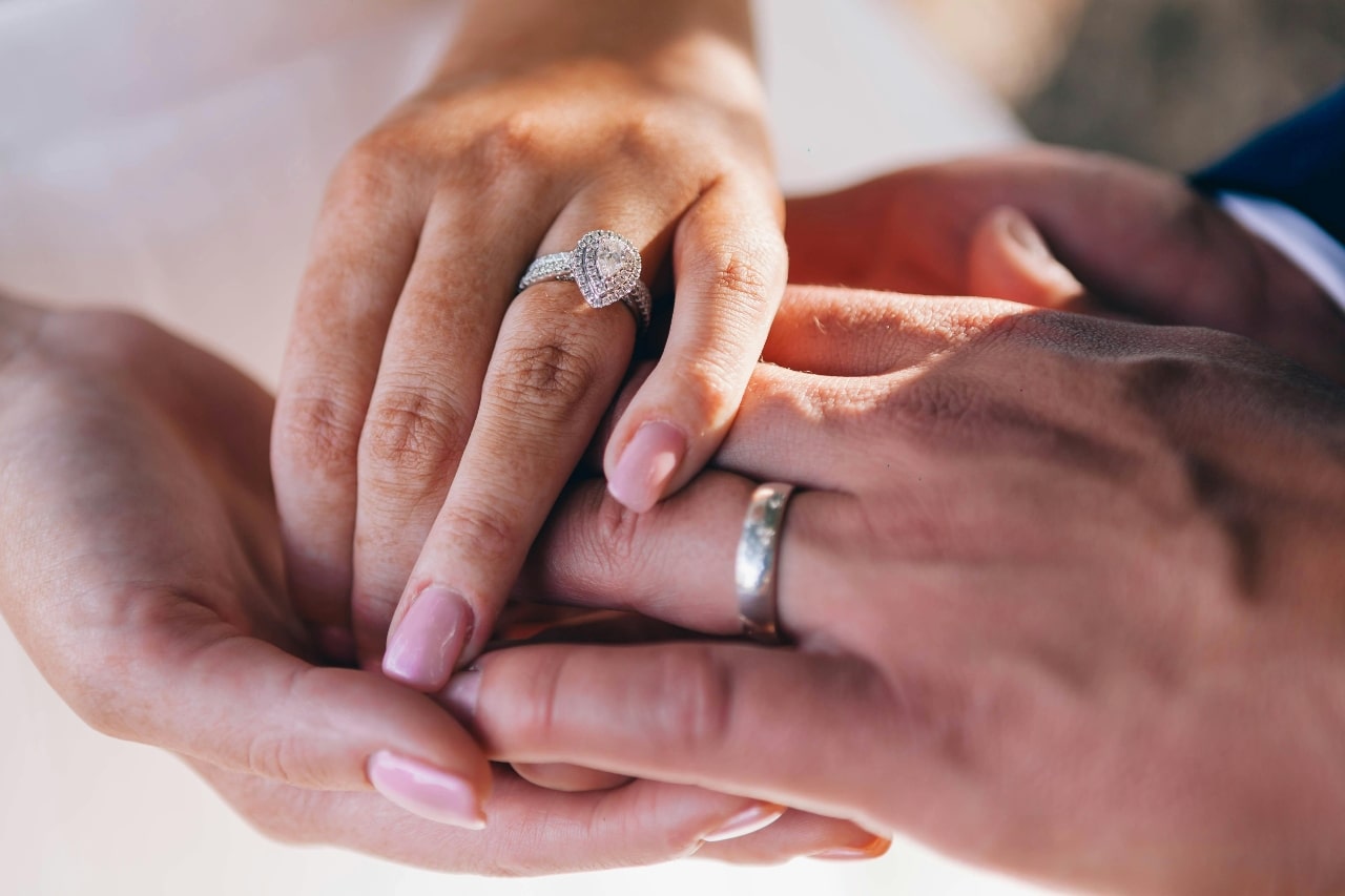 A close-up of a newlywed couple holding hands, with emphasis on his wedding band and her glamorous halo engagement ring.