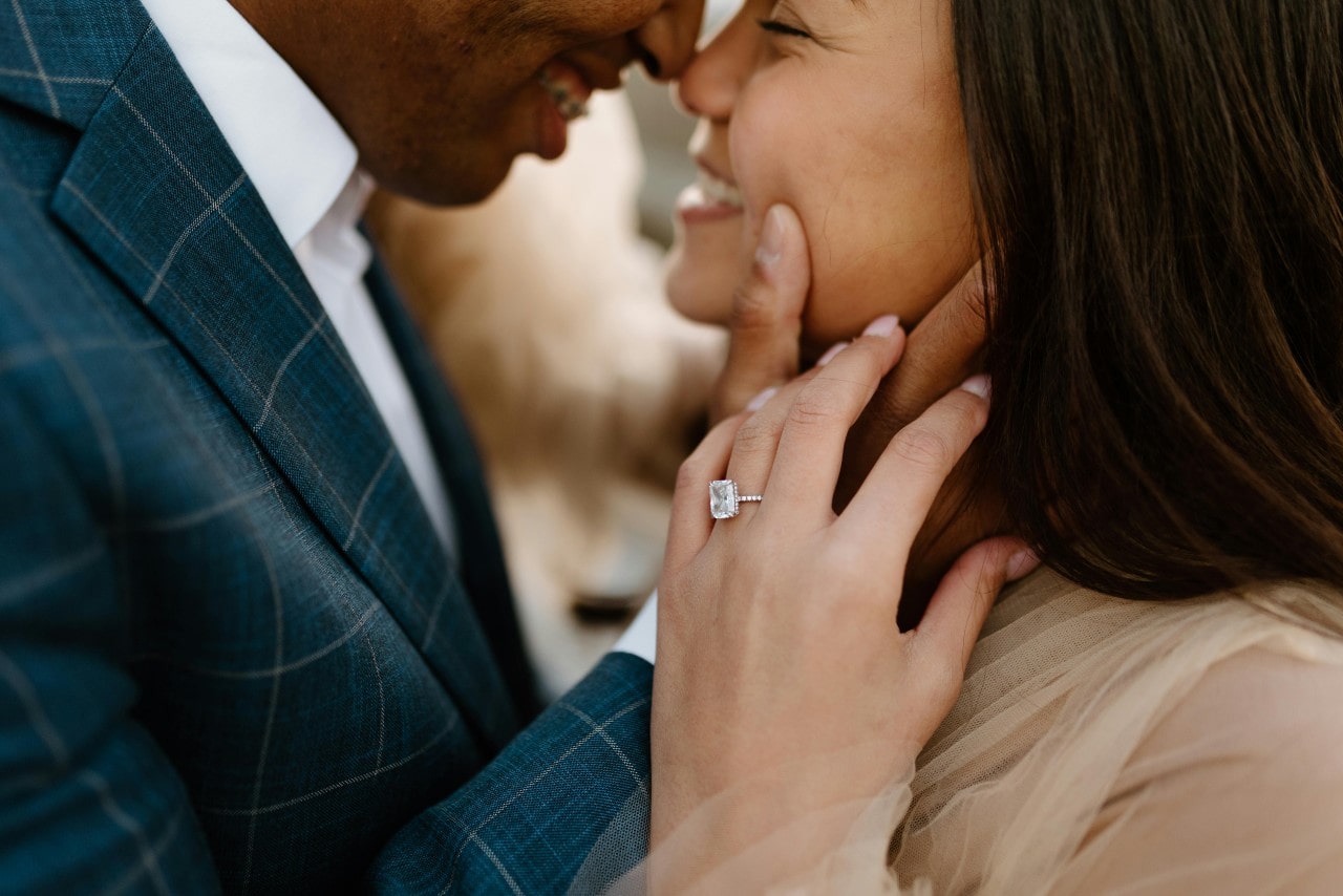A close-up of a happy couple embracing each other, with emphasis on the halo engagement ring on the woman’s finger.
