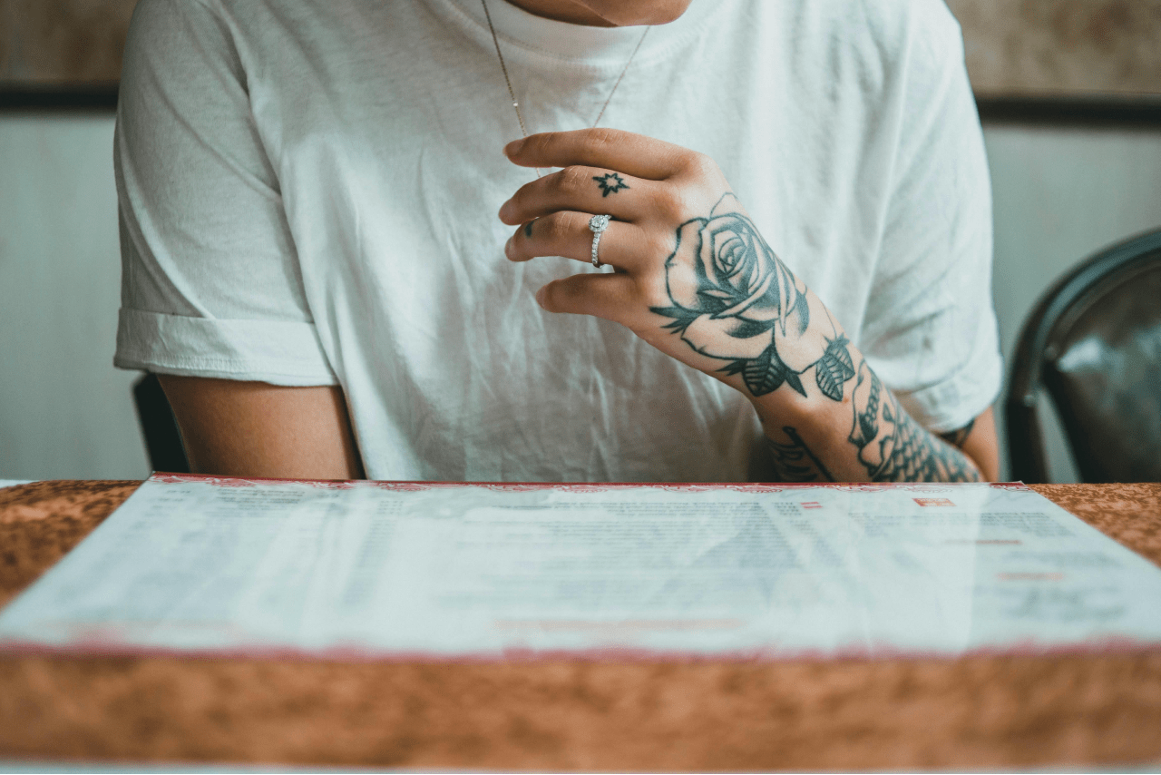 A view of a person’s tattooed arm, an elegant halo engagement ring on their finger, as they peruse a restaurant menu.