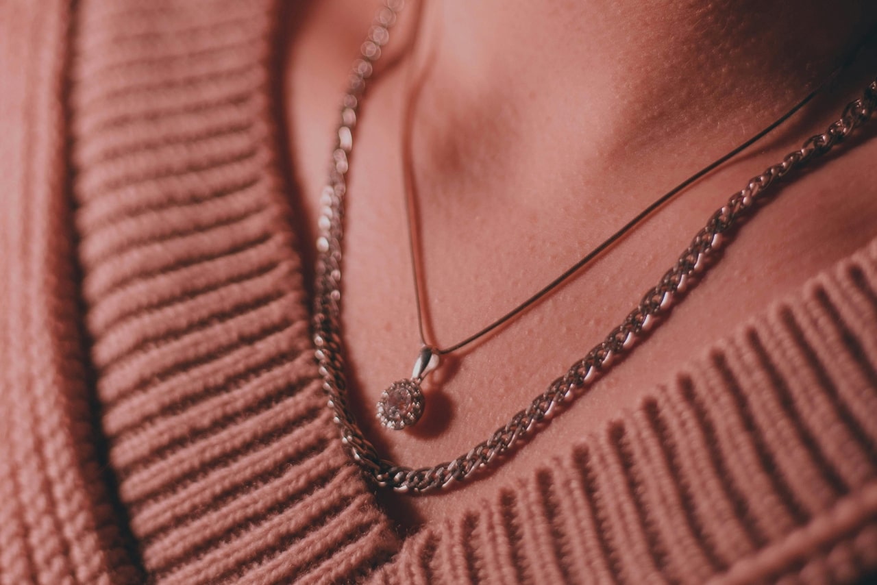 A close-up of a person wearing a diamond halo pendant necklace layered with a thicker chain over a ribbed sweater.