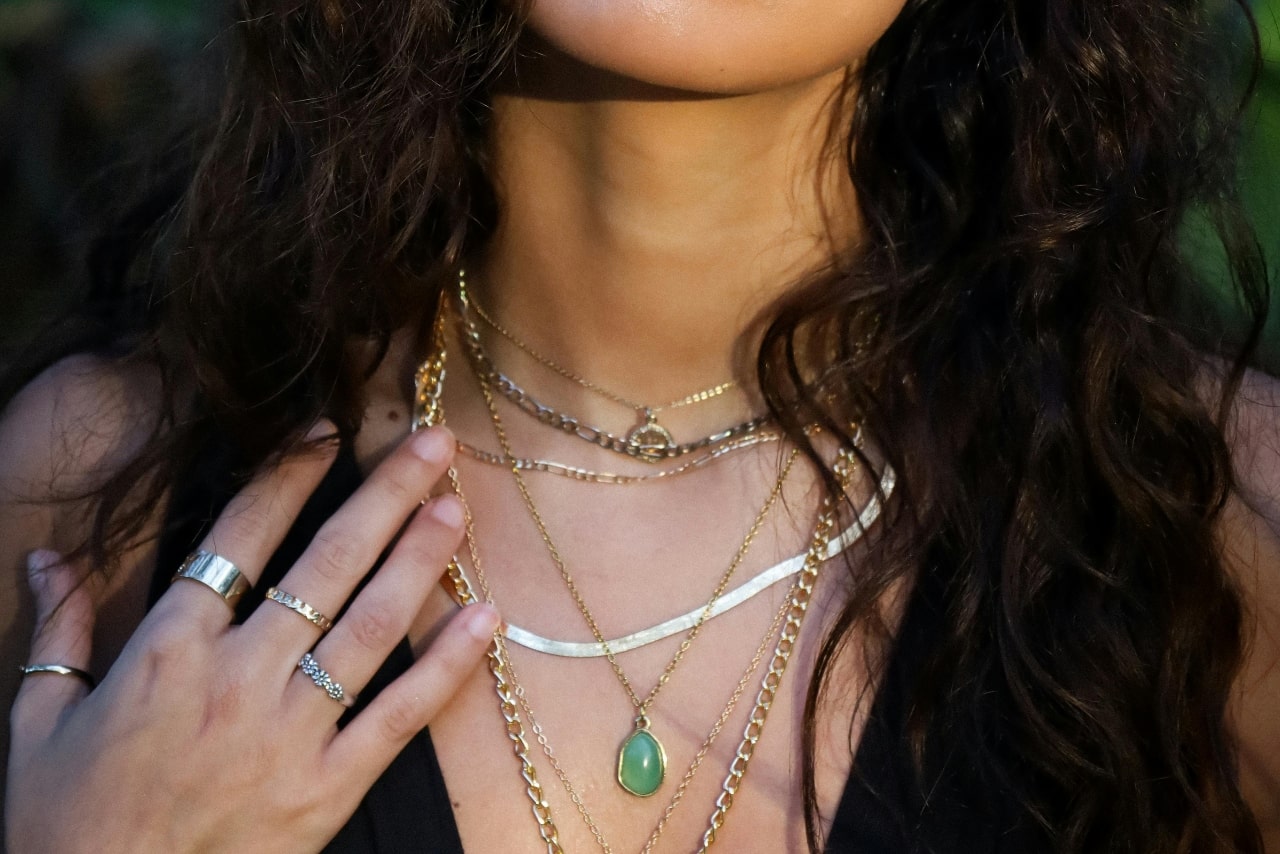 A close-up of a woman wearing multiple layered gold necklaces of varying lengths, including a green teardrop pendant, paired with gold rings and a black top.