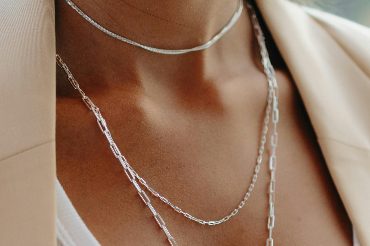A close-up of a woman wearing layered white gold necklaces, including a choker and a longer chain link design, paired with a beige blazer and white t-shirt.