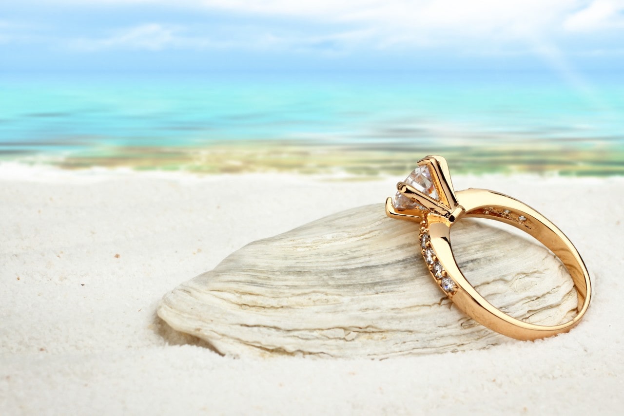 A yellow gold four prong side stone diamond engagement ring on top of the rock in front of the sunny seashore.