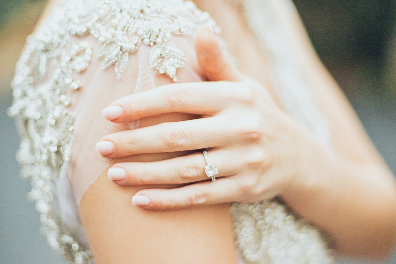 A close up of a bride’s hand adorned with white gold side stone engagement ring on her shoulder.
