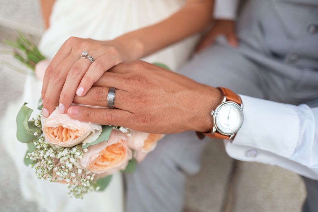 A bride and groom show their wedding rings while holding a bouquet of peach roses and white baby's breath flowers.