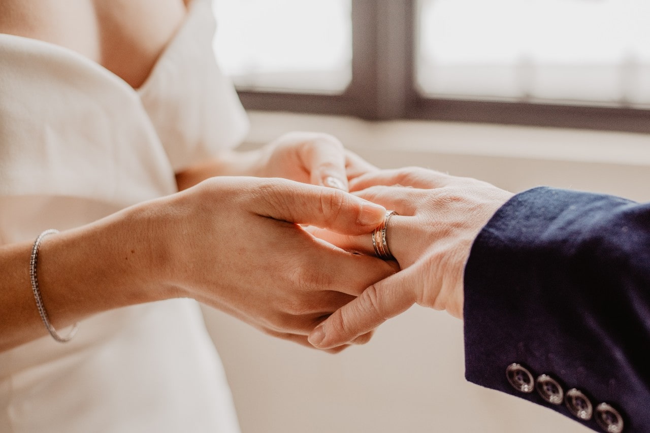 A close up of a bride putting on a wedding band on a man’s hand
