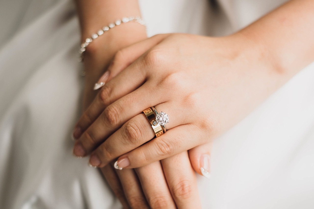 A close up of a yellow gold solitaire diamond engagement ring and matching wedding band on a bride’s hands with manicure nails.