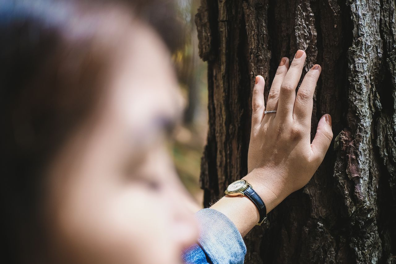A woman wearing a solitaire engagement ring leans against a tree during a hike.