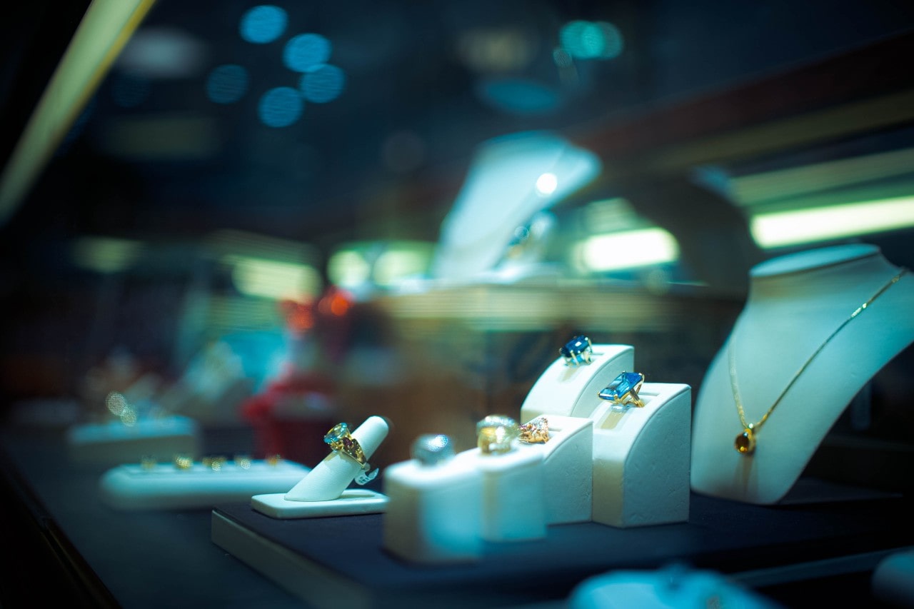 A jewelry display features an array of bold gemstone rings and a gold necklace, set against a high-end boutique background.